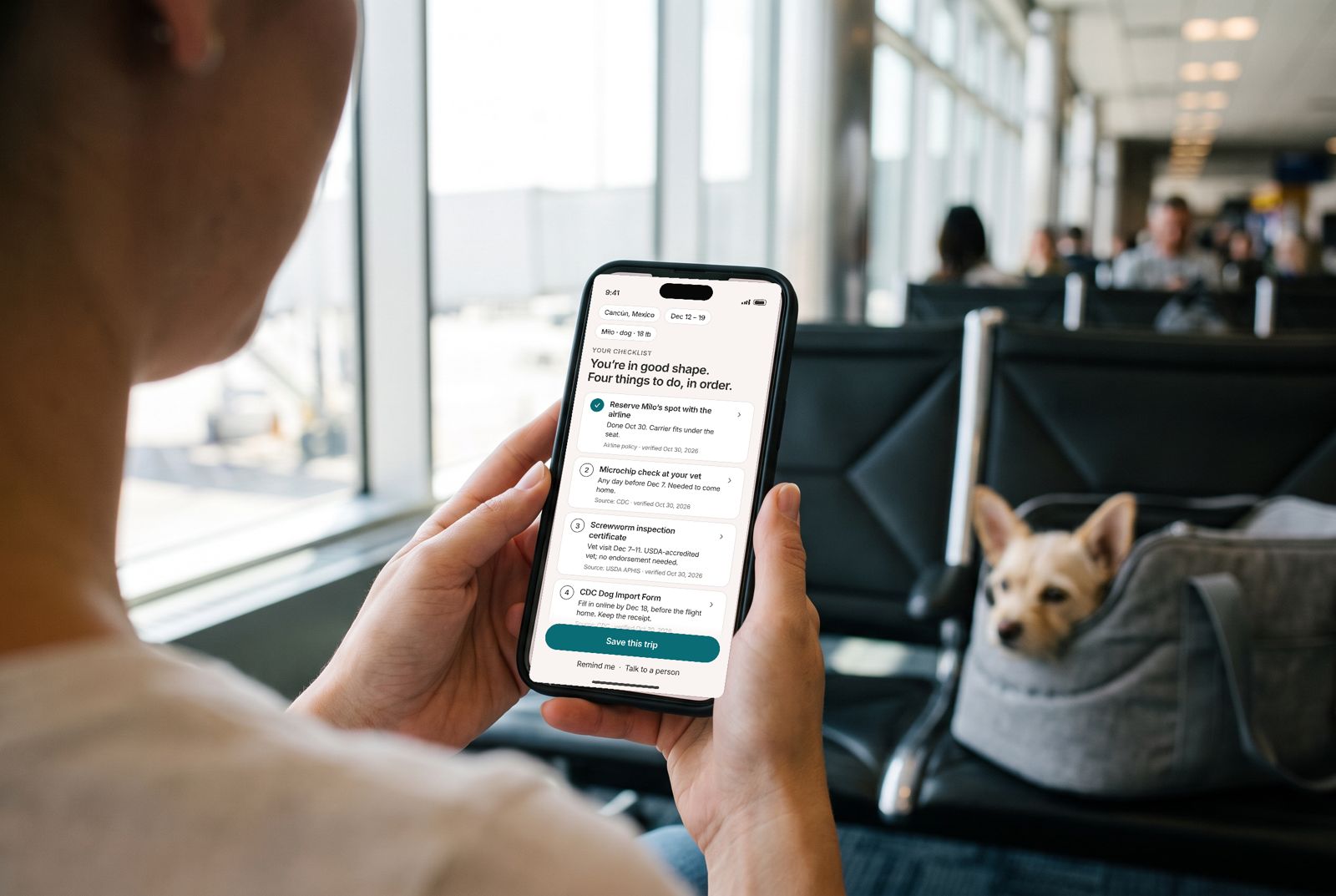 A woman at an airport gate reads a dated checklist on her phone, her small dog in a carrier beside her